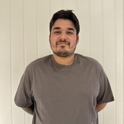 Alexander Dahl, en mann med kort mørkt hår og lett skjegg, smiler forsiktig til kameraet, iført en grå t-skjorte, stående mot en bakgrunn med hvit panel.