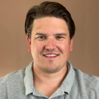 En smilende mann med en vennlig oppførsel, med kort mørkt hår og en uformell grå poloskjorte, satt mot et nøytralt bakteppe.