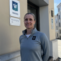 En kvinne som smiler utendørs nær en bygningsinngang med et skilt som leser "Wang" og en pil som peker mot venstre merket "Elevinngang." Hun har på seg en grå sporty jakke med logo.