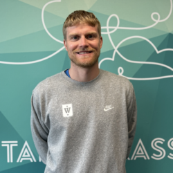 Emil Jonassen, en mann med kort blondt hår og skjegg, iført en grå Nike-genser, står smilende foran en blågrønn vegg med hvit, abstrakt strek og delvis synlig tekst.